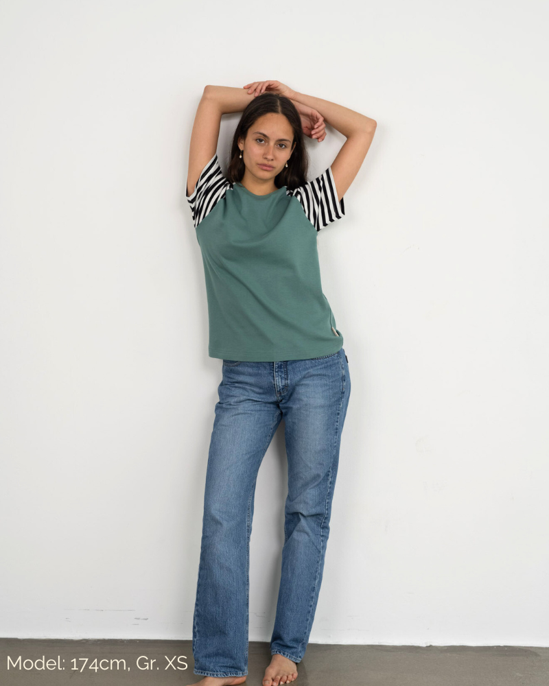 Eine Frau lehnt lässig an der Wand und trägt das Raglan Shirt in Grün mit gestreiften Ärmeln der Marke Oktopulli