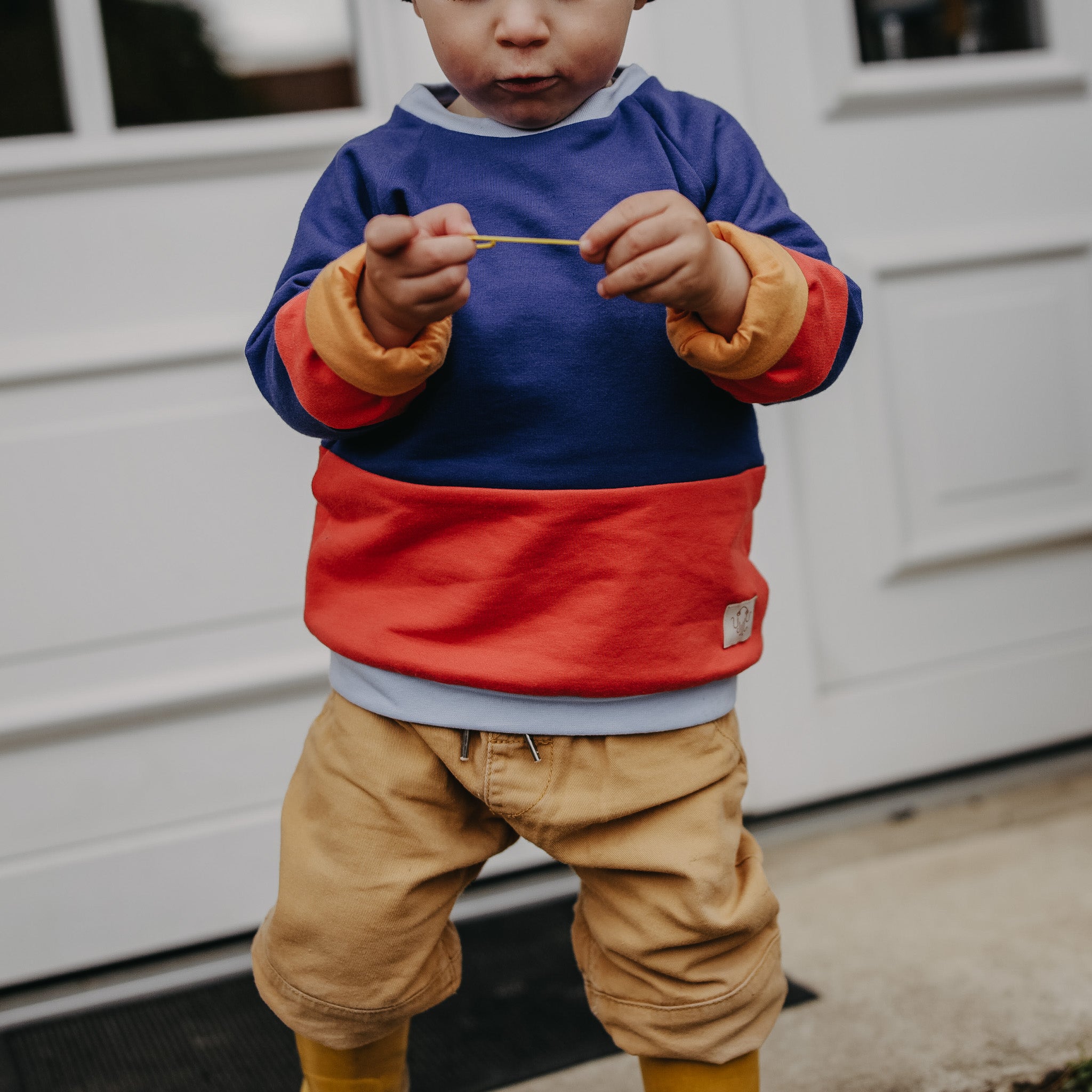 Kleinkind steht vor einer weißen Tür. Gesicht ist ab dem Mund nach oben hin vom Foto abgeschnitten. Hält einen kleinen Stock in der Hand. Kind trägt einen orange blauen colorblocking Pullover und eine kurze beige Hose.  .