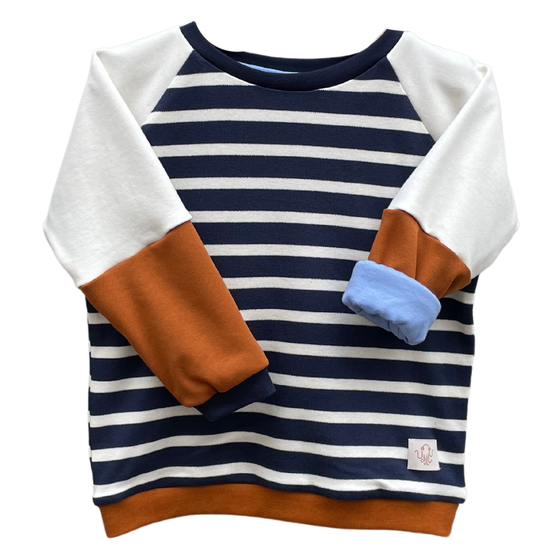 Kinderpulli Eike mit Raglanärmeln. Korpus marineblau und weiß gestreift. Ärmel zweiteilig. Oberer Farbe cremefarben, unterer Farbe kaffeebraun. Halsbündchen und Ärmelbündchen in marineblau, Saumbündchen kaffeebraun. Ein Ärmel ist gekrempelt und zeigt ein hellblaues Futter.