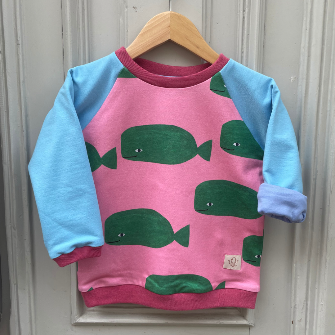 Beispielfoto: Raglanpulli für Kinder. Körper pink mit dunkelgrünen gezeichneten Walen. Ärmel taubenblau. Innenfutter an den Ärmeln dunkleres Taubenblau. Bündchen Pink meliert.  