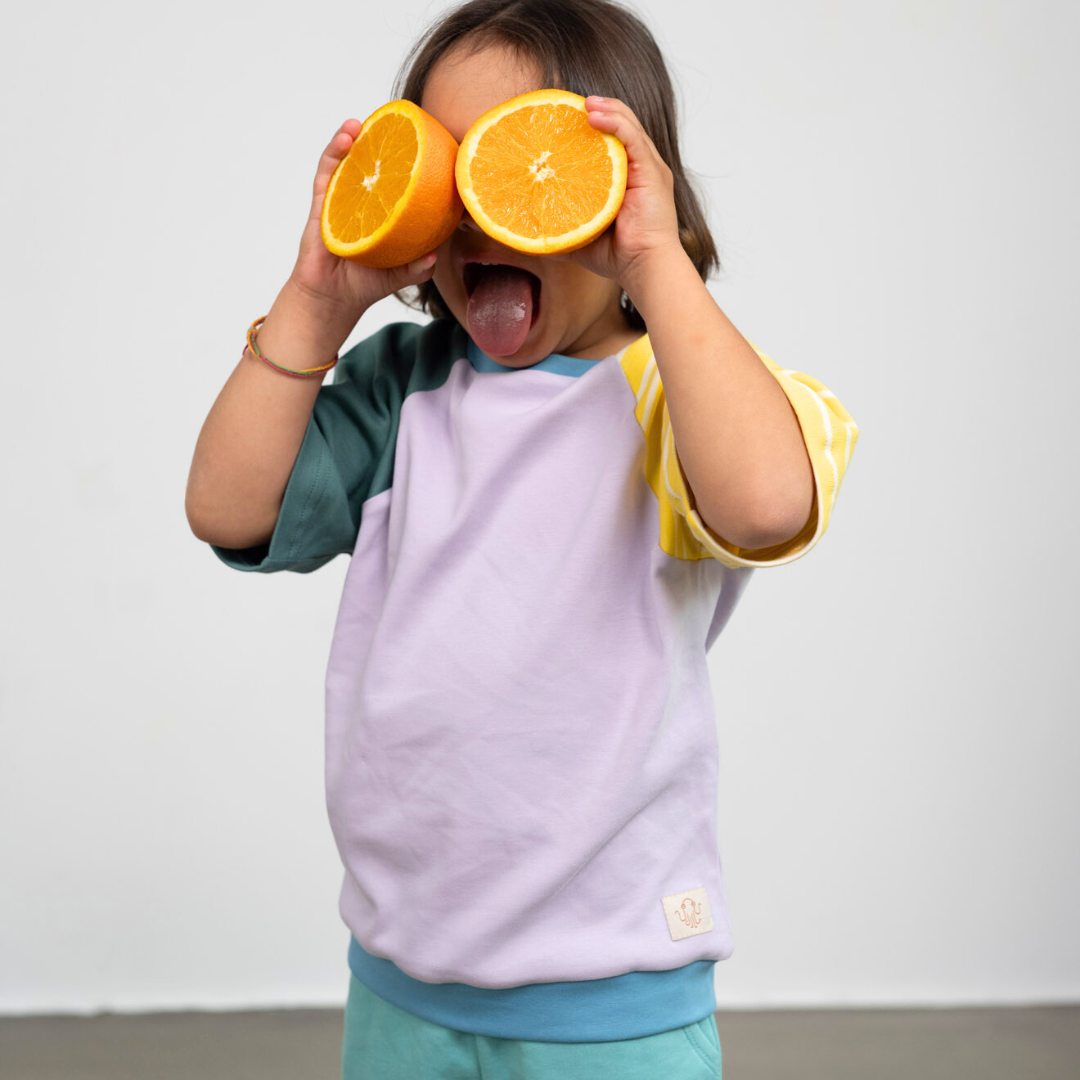 Ein Kind trägt das faire Shirt von Oktopulli aus Bio-Baumwolle in Flieder und hält sich Orangen vor die Augen