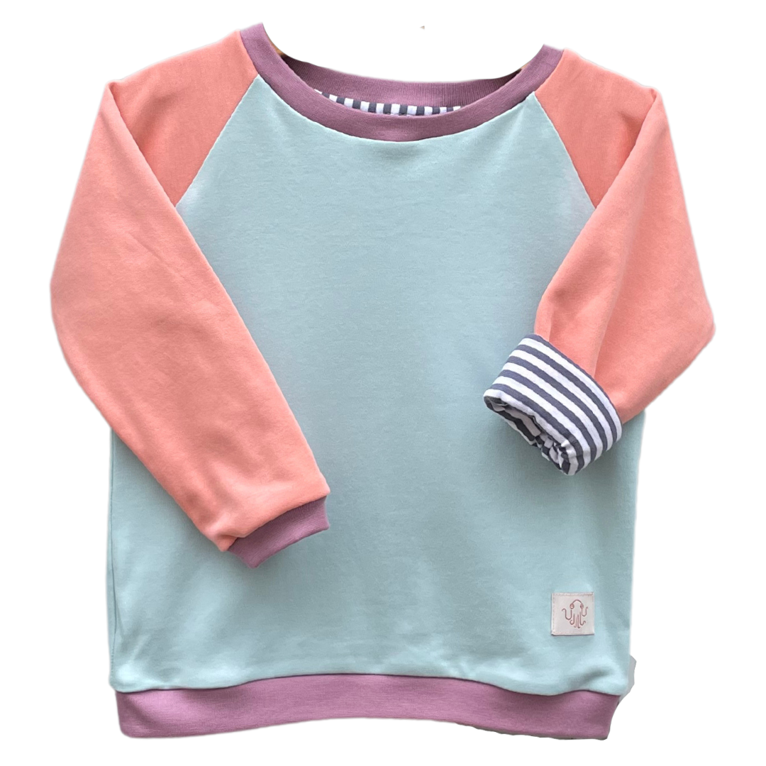 Kinderpulli Kaya mit Raglanärmeln. Korpus hellblau. Ärmel rosa. Halsbündchen, Ärmelbündchen und Saumbündchen altrosa. Ein Ärmel ist gekrempelt und zeigt ein gestreiftes Futter mit dunkelblauen und weißen gleich großen Streifen. 