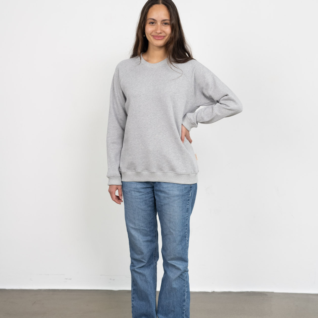 Eine Frau steht lächelnd vor einer Wand und trägt den genderneutralen Kuschelpullover in Grau aus Bio-Baumwolle von Oktopulli