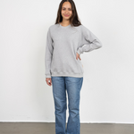 Eine Frau steht lächelnd vor einer Wand und trägt den genderneutralen Kuschelpullover in Grau aus Bio-Baumwolle von Oktopulli