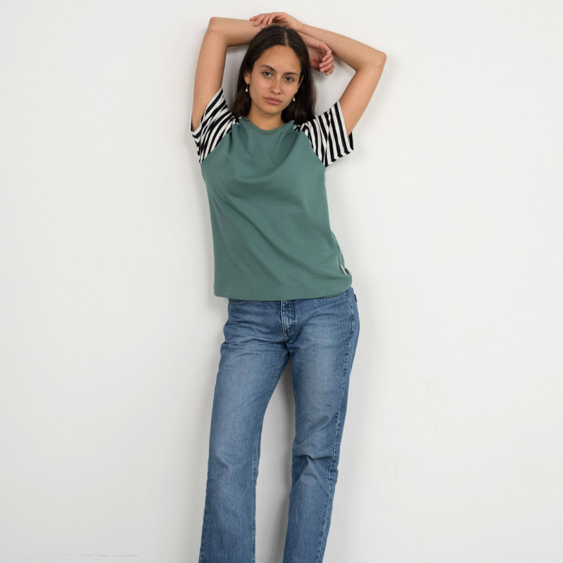 Eine Frau lehnt lässig an der Wand und trägt das Raglan Shirt in Grün mit gestreiften Ärmeln der Marke Oktopulli