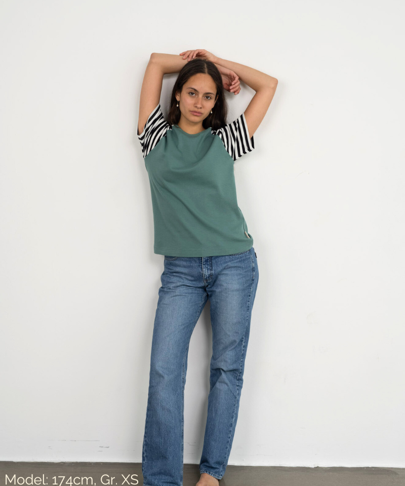 Eine Frau lehnt lässig an der Wand und trägt das Raglan Shirt in Grün mit gestreiften Ärmeln der Marke Oktopulli