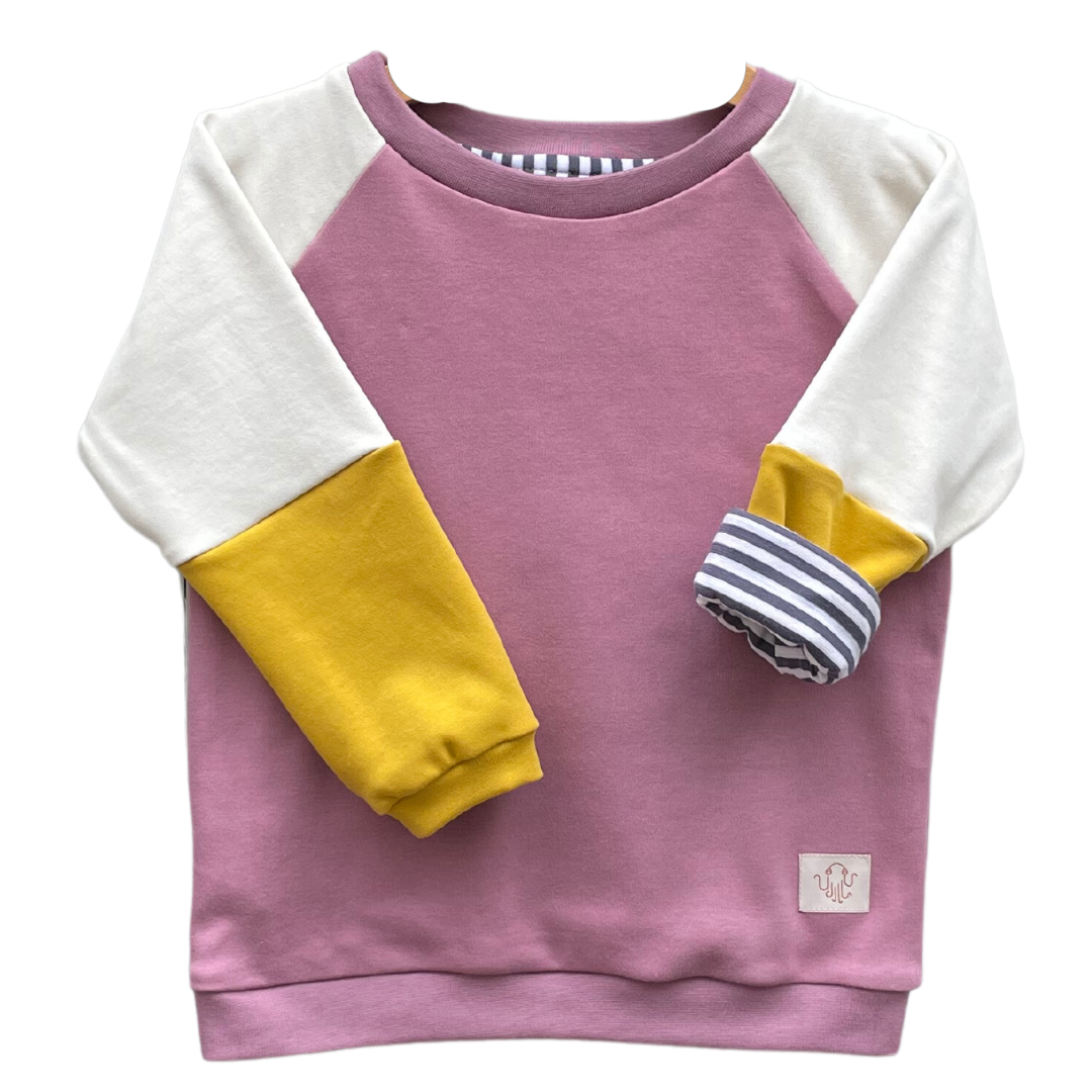 Kinderpulli Benja mit Raglanärmeln. Korpus ist altrosa. Ärmel zweiteilig. Obere Farbe ist cremefarben, unterer Teil ist gelb. Halsbündchen und Saumbündchen in altrosa, Ärmelbündchen in gelb. Ein Ärmel ist gekrempelt und zeigt ein gestreiftes Futter mit dunkelblauen und weißen gleich großen Streifen. 