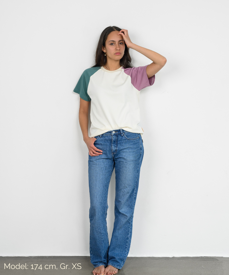 Eine Frau trägt ein weißes Raglan Shirt mit bunten Ärmeln der Marke Oktopulli im Unisex-Schnitt und lehnt an einer Wand