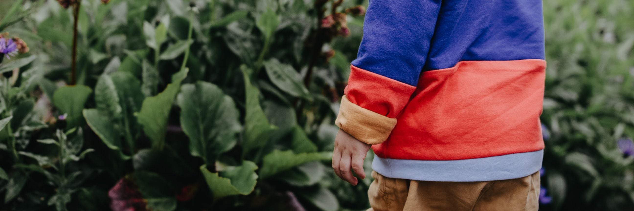 Nahaufnahme von einem Kind im Garten, das einen Oktopulli im Color-Blocking Design trägt. Die obere Hälfte des Pullis ist blau, die untere orange-rot. Der Ärmel ist umgeschlagen, wodurch das senfgelbe Innenfutter sichtbar wird.