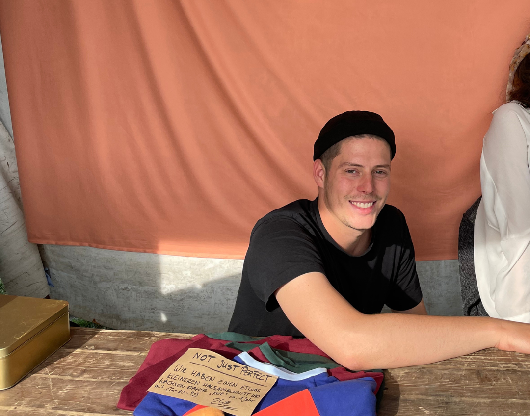 Portrait von dem Mitarbeiter Bene. Er sitzt hinter einem Marktstand und lacht freundlich in die Kamera.