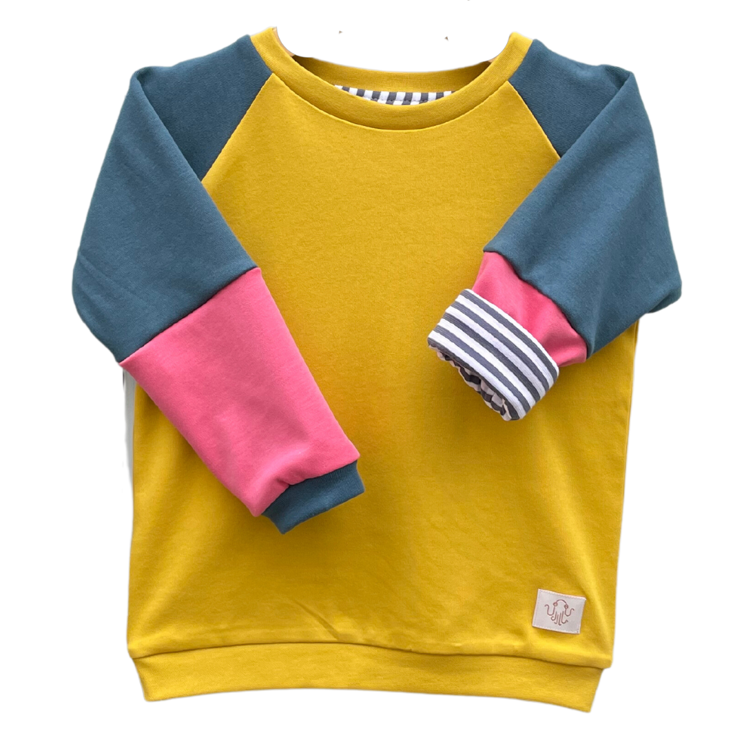 Kinderpulli Evan mit Raglanärmeln. Korpus gelb. Ärmel zweiteilig. Obere Farbe dunkelgrün, untere Farbe magenta. Halsbündchen und Ärmelbündchen dunkelgrün, Saumbündchen gelb. Ein Ärmel ist umgekrempelt und zeigt ein gestreiftes Futter mit dunkelblauen und weißen gleich großen Streifen. 
