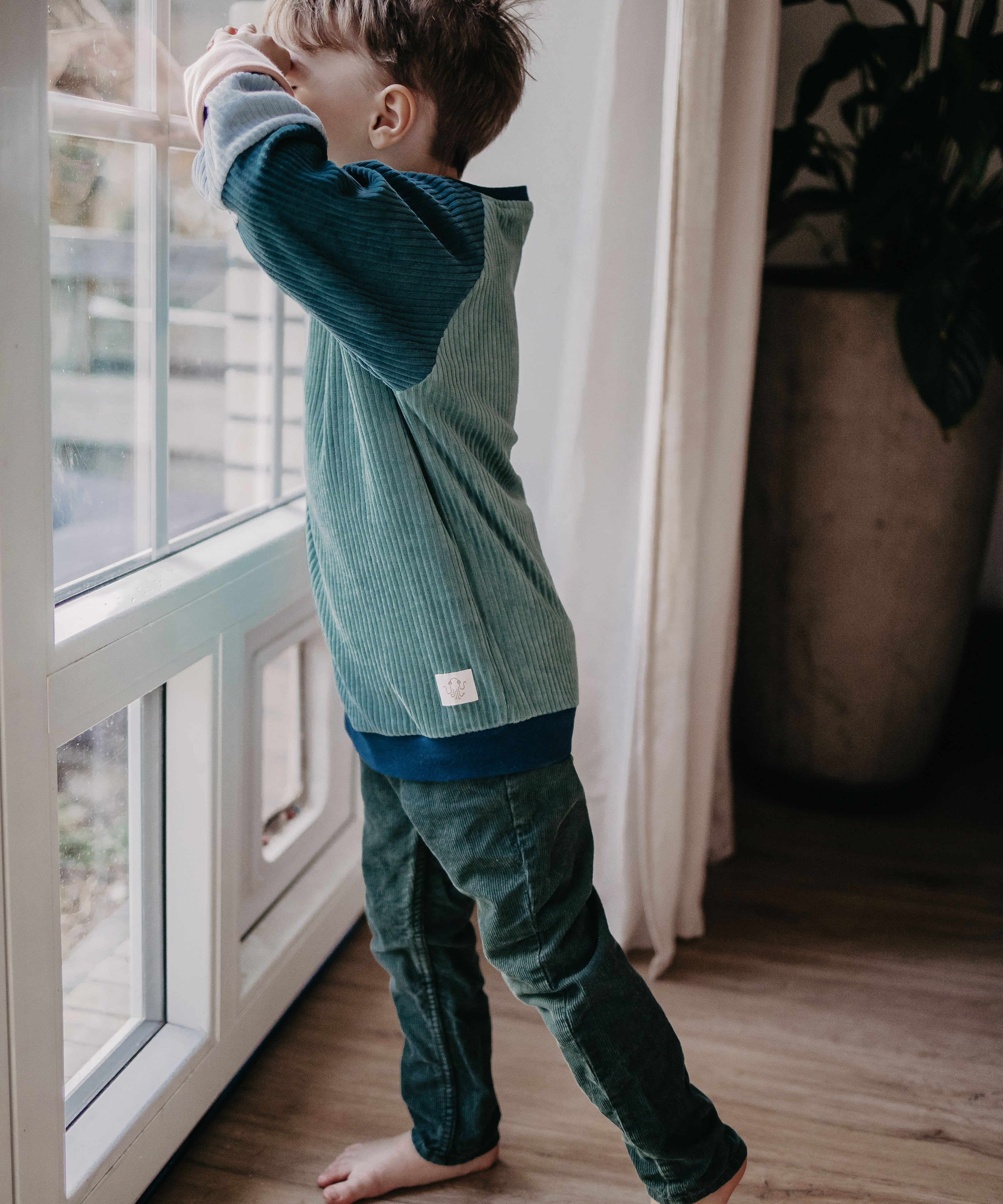  Ein Kind guckt aus dem Fenster und trägt dabei den blauen Alltagsbegleiter im Unisex-Schnitt von Oktopulli