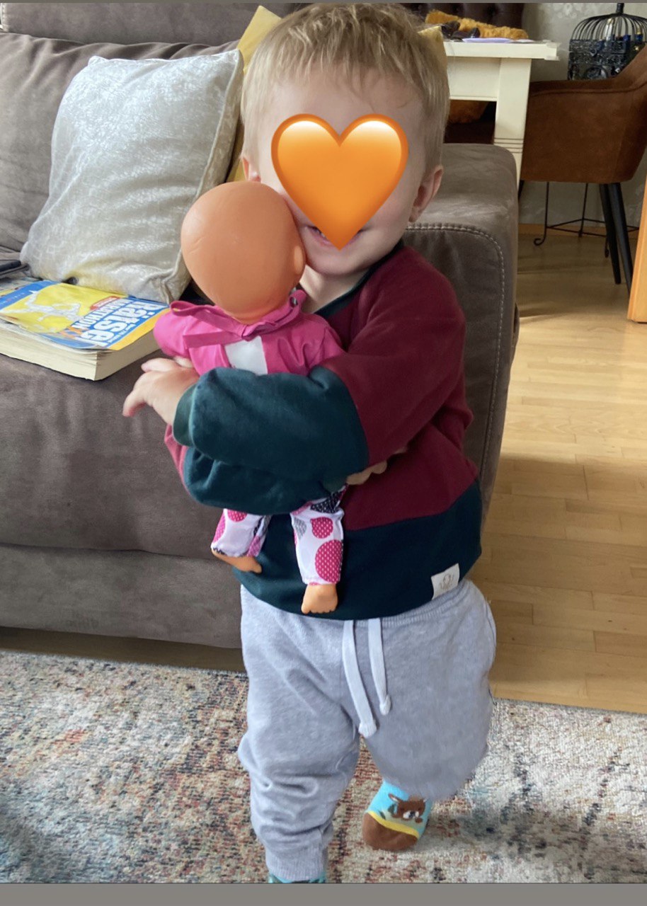 Kind steht vor einer Couch, trägt einen grün-roten Oktopulli und hat eine Puppe im Arm. Das Gesicht des Kindes ist mit einem Herz Emoji verdeckt.