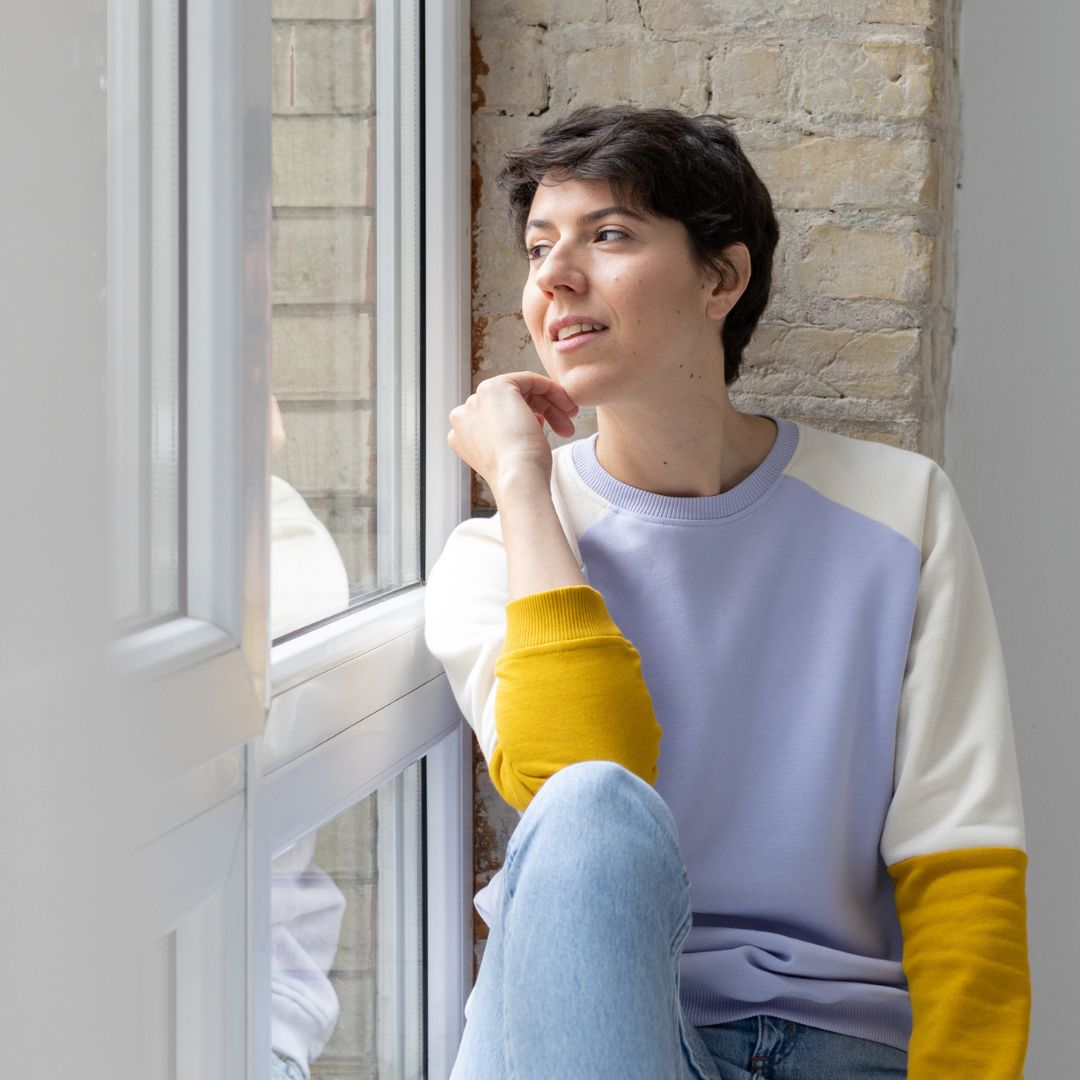 Eine Frau sitzt am Fenster und schaut hinaus. Sie trägt einen dreifarbigen Oktopulli mit fliederfarbenen Körper, weißen Oberarmen und senfgelben Unterarmen.
